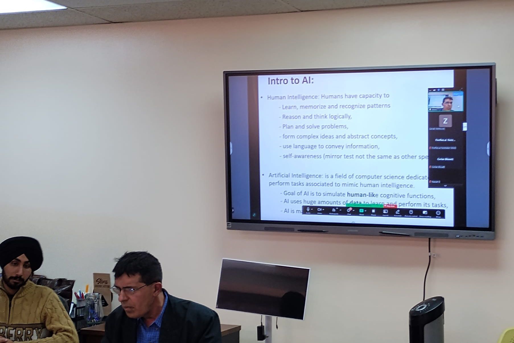 A presentation on a screen with people in the room, including two men in the foreground.