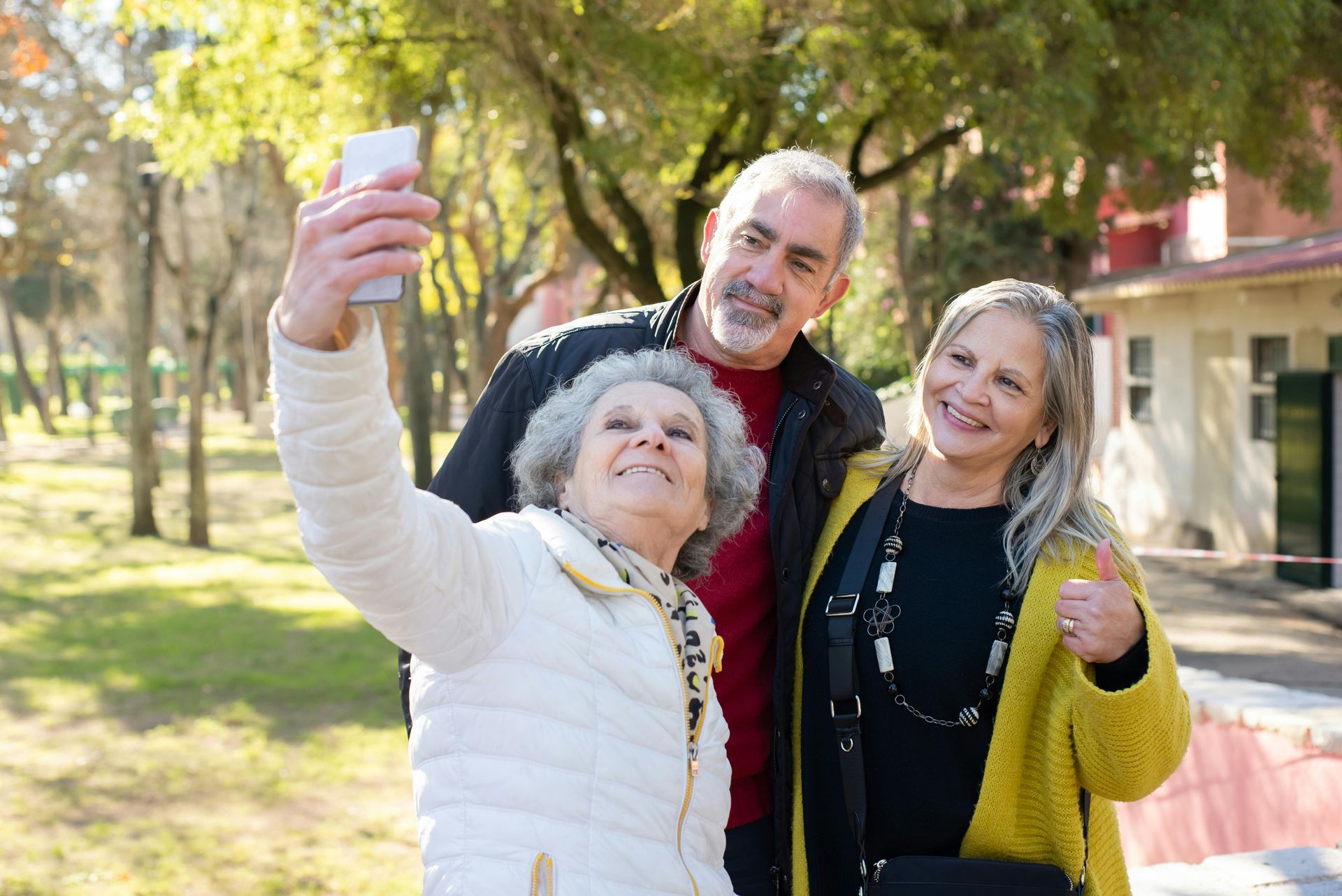 Three older adults smile while taking a selfie outdoors in a park.