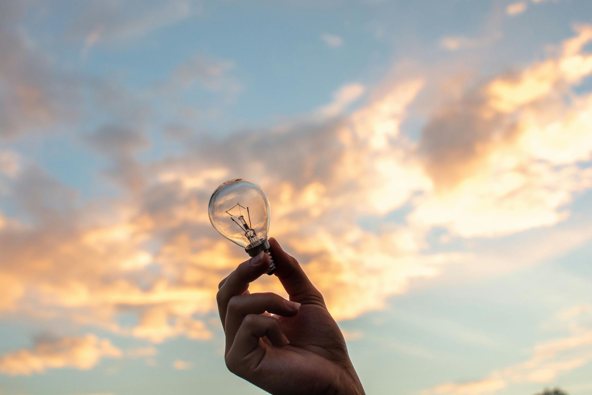 A person is holding a light bulb in their hand in front of a cloudy sky.