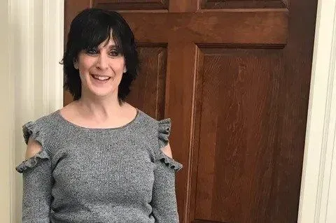Woman in gray sweater with shoulder cutouts smiling in front of a wooden door.