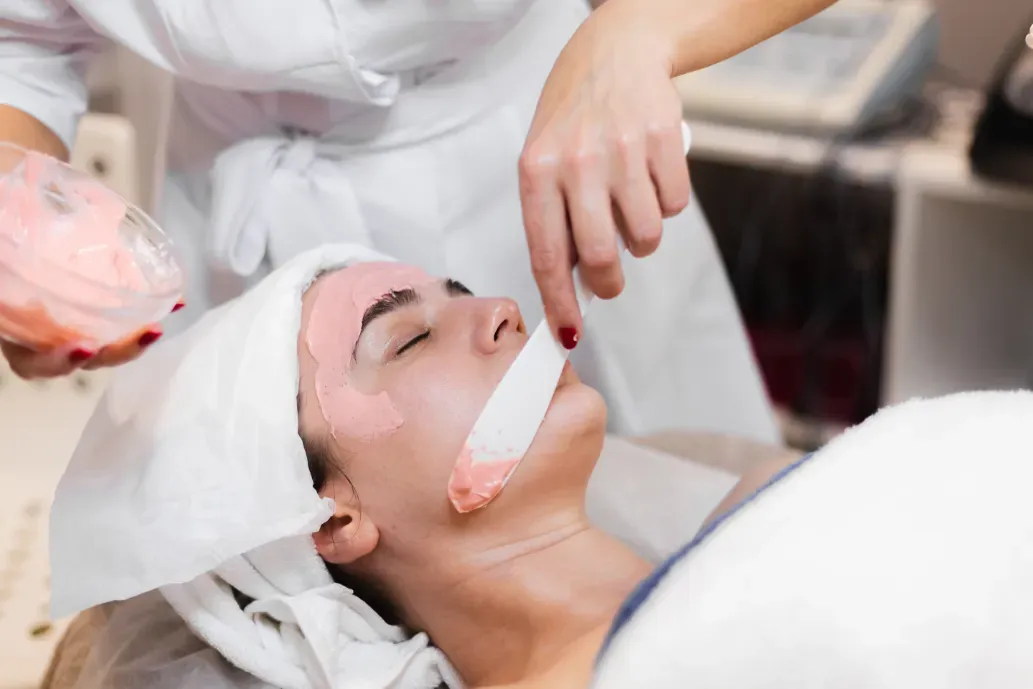Woman receiving facial treatment; pink mask applied to her face by a gloved hand in a salon setting.