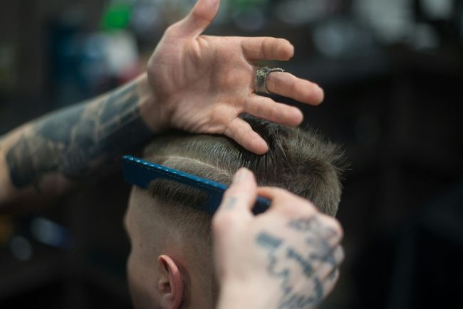 Barber combing hair, shaping a fade. Hands with tattoos; dark salon setting.