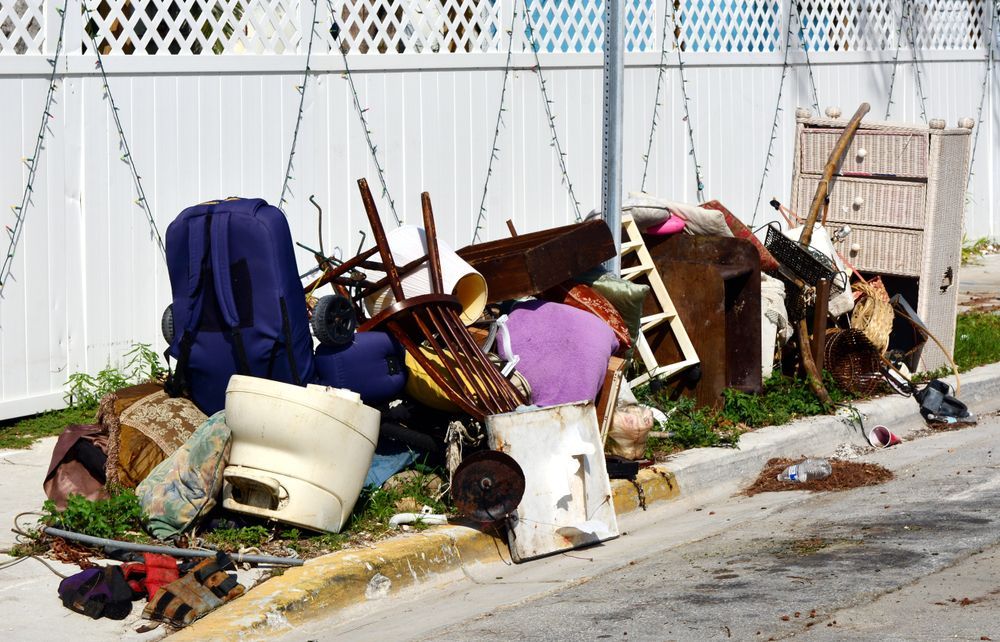 Pile of discarded furniture and trash on a curb near a white fence.