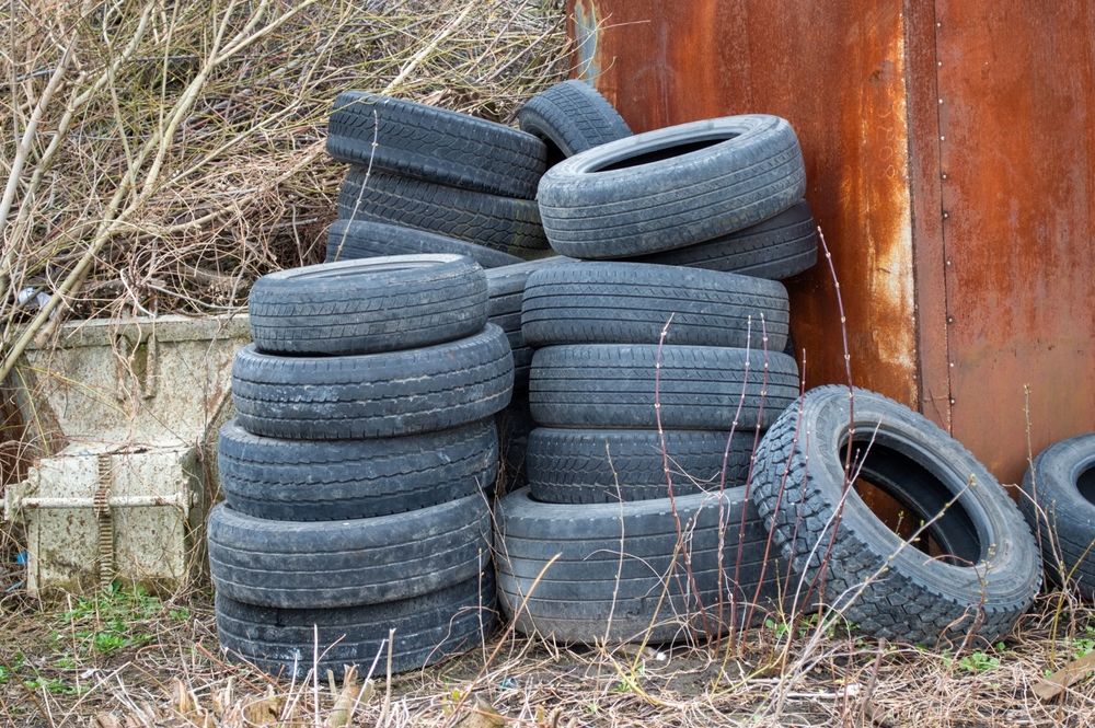 Piles of used tires leaning against a rusty metal wall, surrounded by dry grass and weeds.