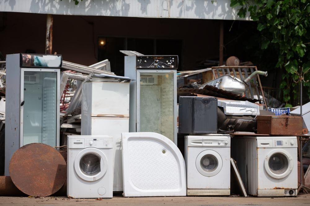 Junked appliances and metal items piled outdoors; includes refrigerators, washers, and a shower base.