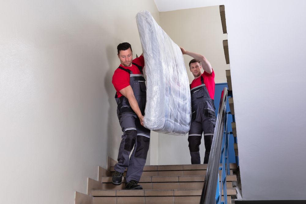 Two people in red shirts and gray overalls carrying a wrapped mattress up stairs.