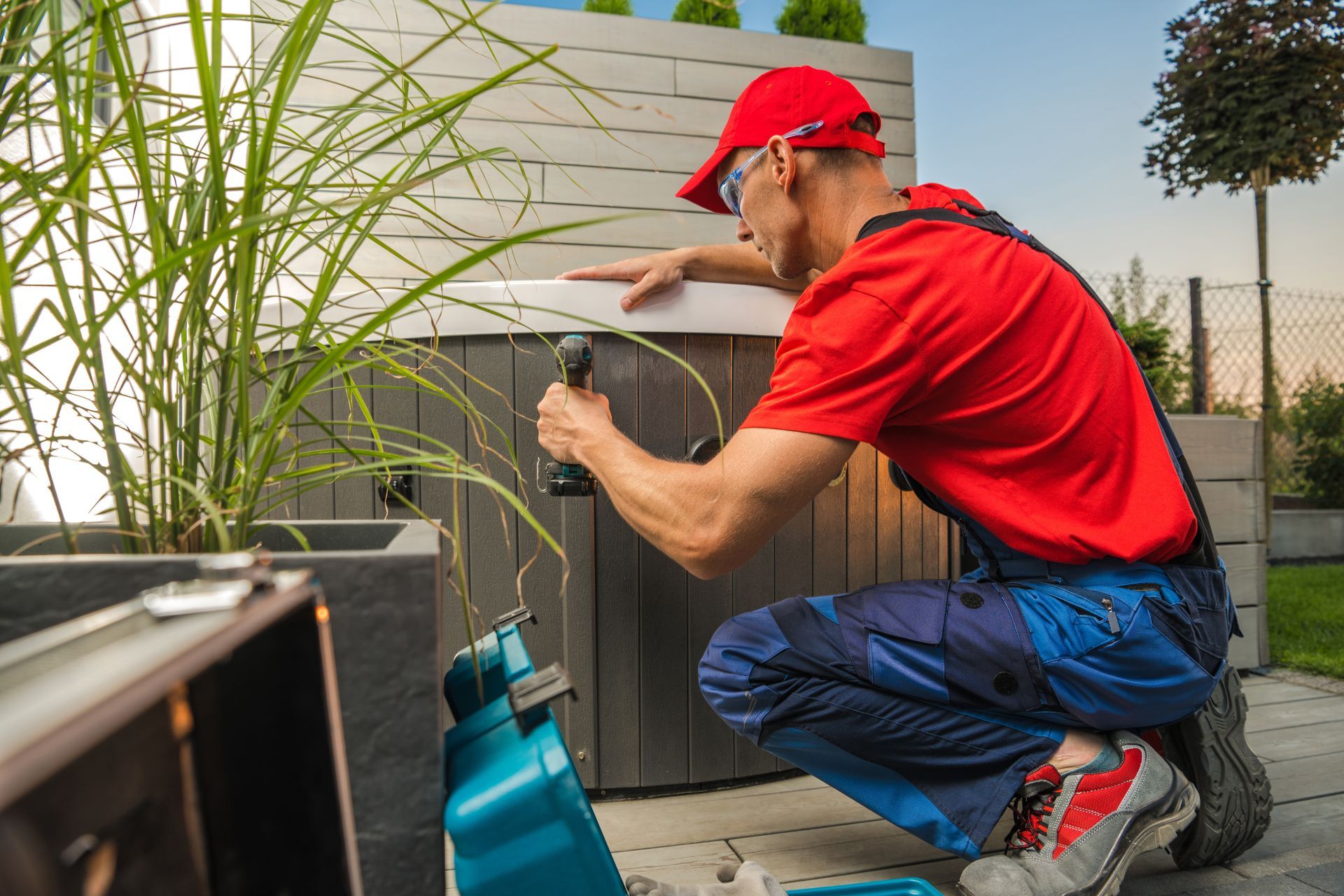 Man in red shirt and blue overalls using a drill outdoors on a gray structure, near a plant.