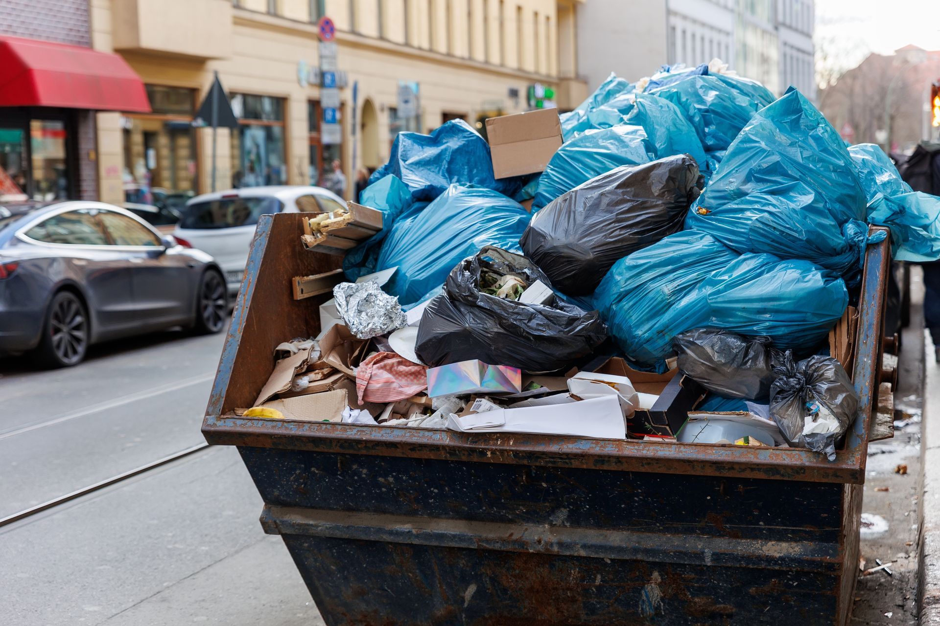Overflowing trash bin on a city street, filled with blue and black trash bags and loose waste.