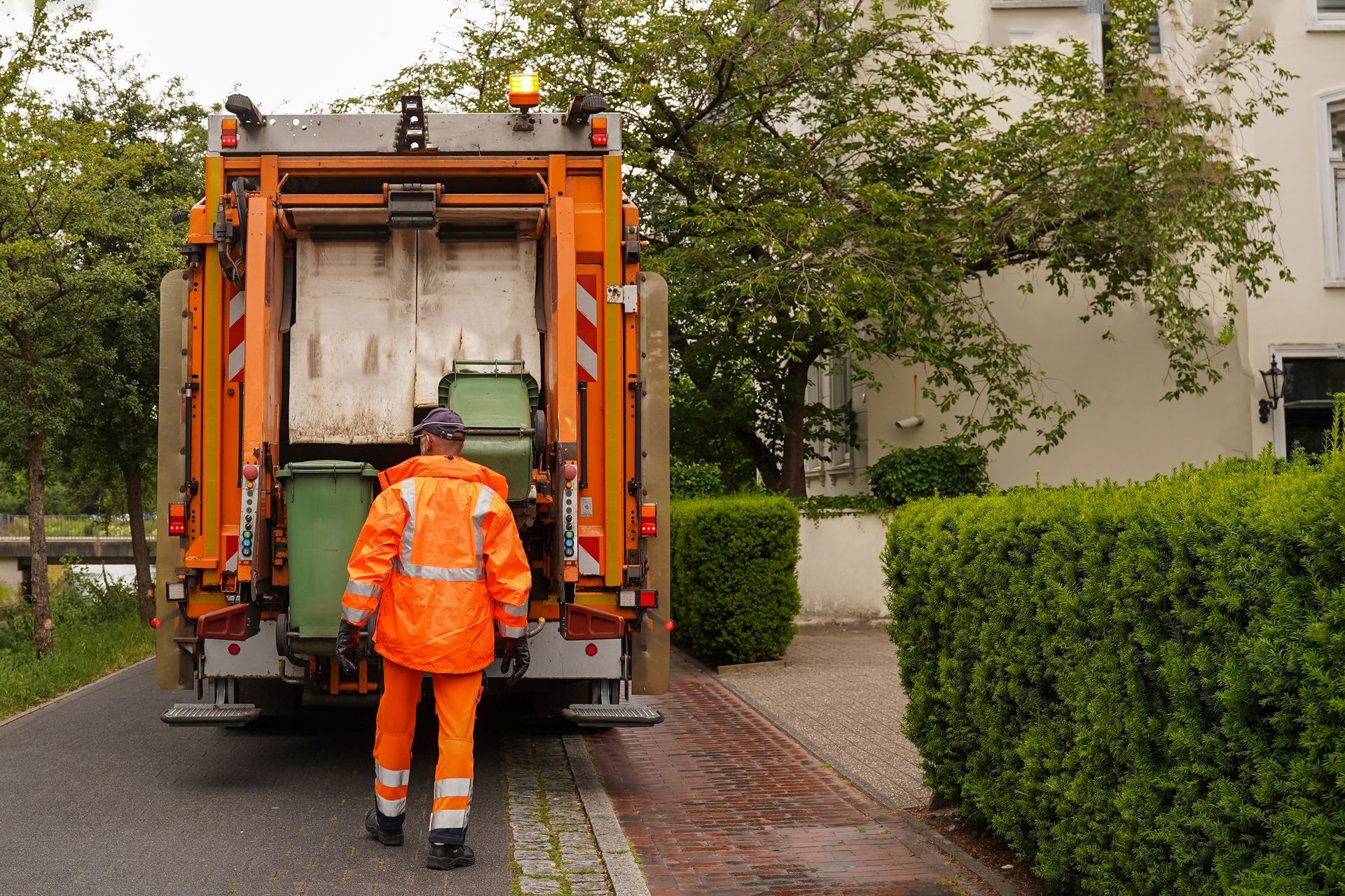 Garbage truck worker in orange suit, loading bins on a residential street.