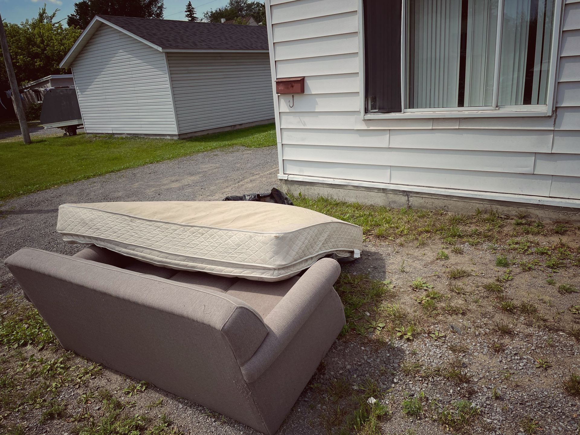 Curb-side junk: mattress and sofa leaning on each other on a gravel patch, near a white house and garage.
