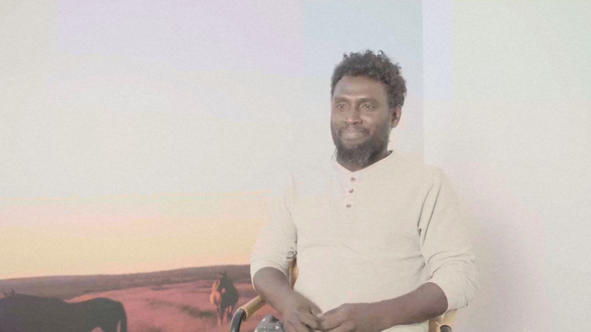 A man with a beard is sitting in a chair in front of a painting.