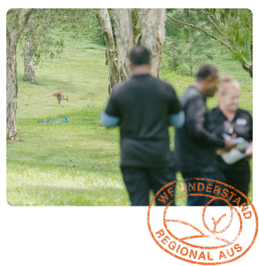 A group of workers in black shirts stand in the foreground, while a kangaroo hops across a grassy area with scattered trees.