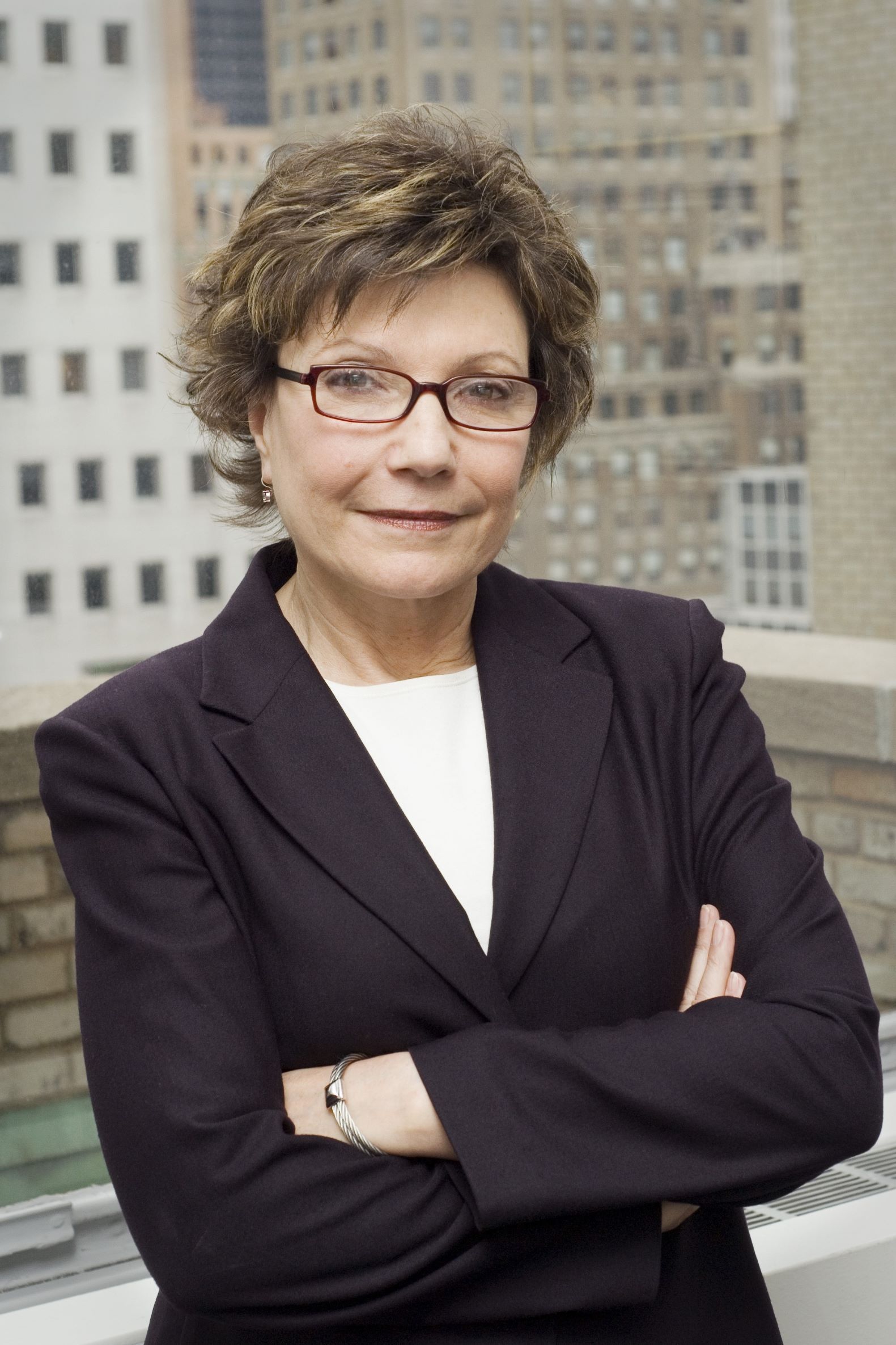 A professional standing with arms crossed, wearing a dark blazer and glasses in front of a blurred city building backdrop.