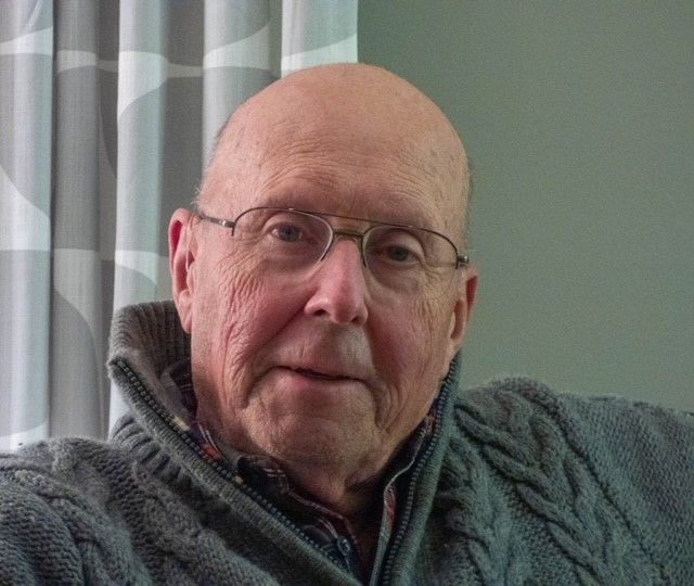 A bald man wearing glasses and a grey sweater is sitting on a couch.