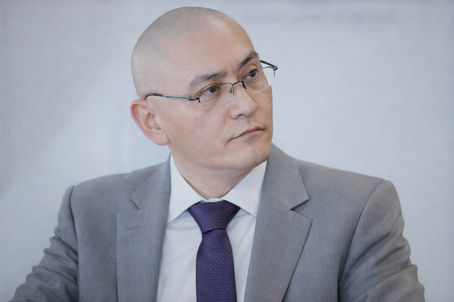 A person in a grey suit and purple tie wears glasses and looks off-camera against a plain, light-colored background.