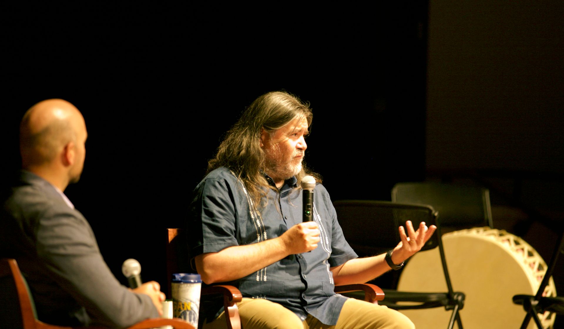 Two men are sitting in chairs on a stage talking into microphones.