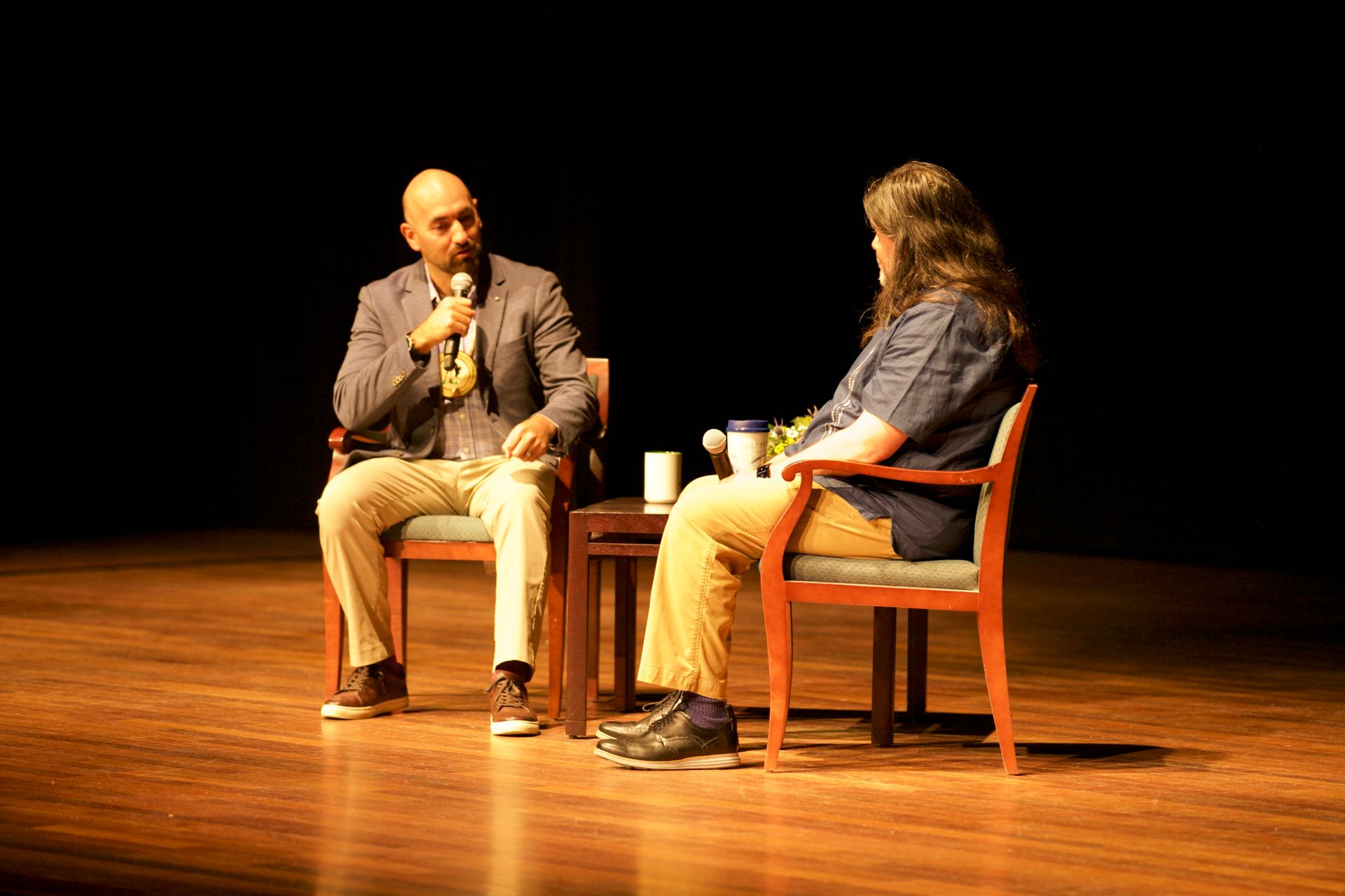 Two men are sitting in chairs on a stage talking to each other.