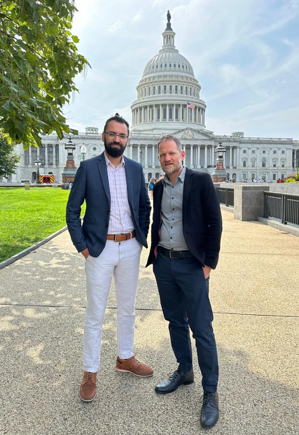 Two men in suits stand in front of the US Capitol Building. One wears white pants, the other navy.