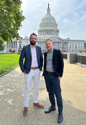 Two men in suits stand in front of the US Capitol Building. One wears white pants, the other navy.