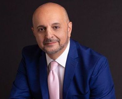 A man in a blue suit and pink tie is sitting on a stool.