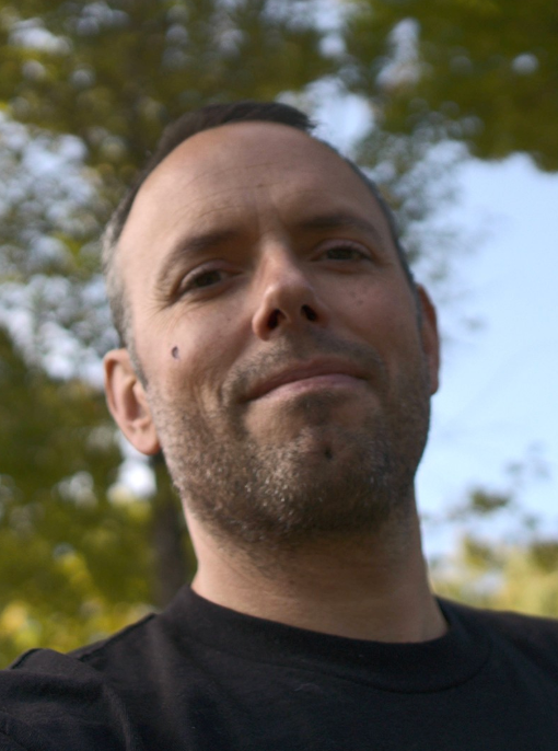 A person with short hair and facial stubble smiling slightly outdoors, with blurred trees in the background.