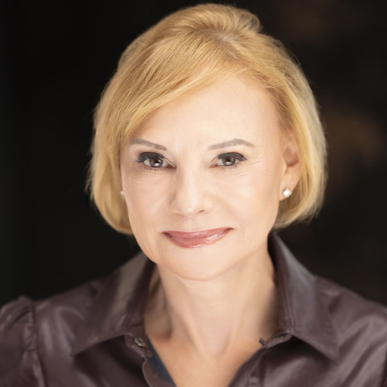 Blonde woman in brown shirt, smiling, looking at the camera against a dark background.