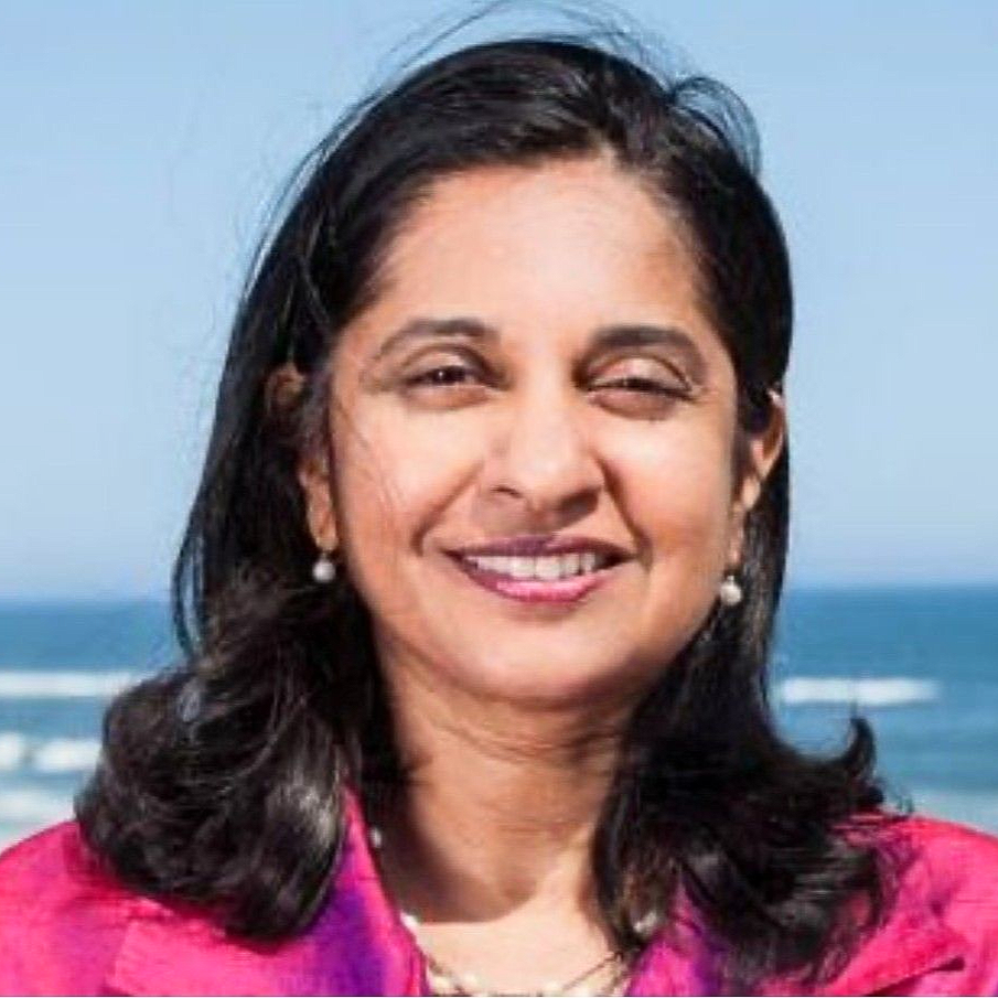 Woman with glasses wearing a turquoise blazer and pearl necklace, smiling.