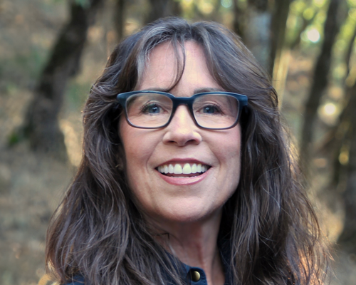 Woman smiling, wearing glasses, outdoors in front of blurred trees.