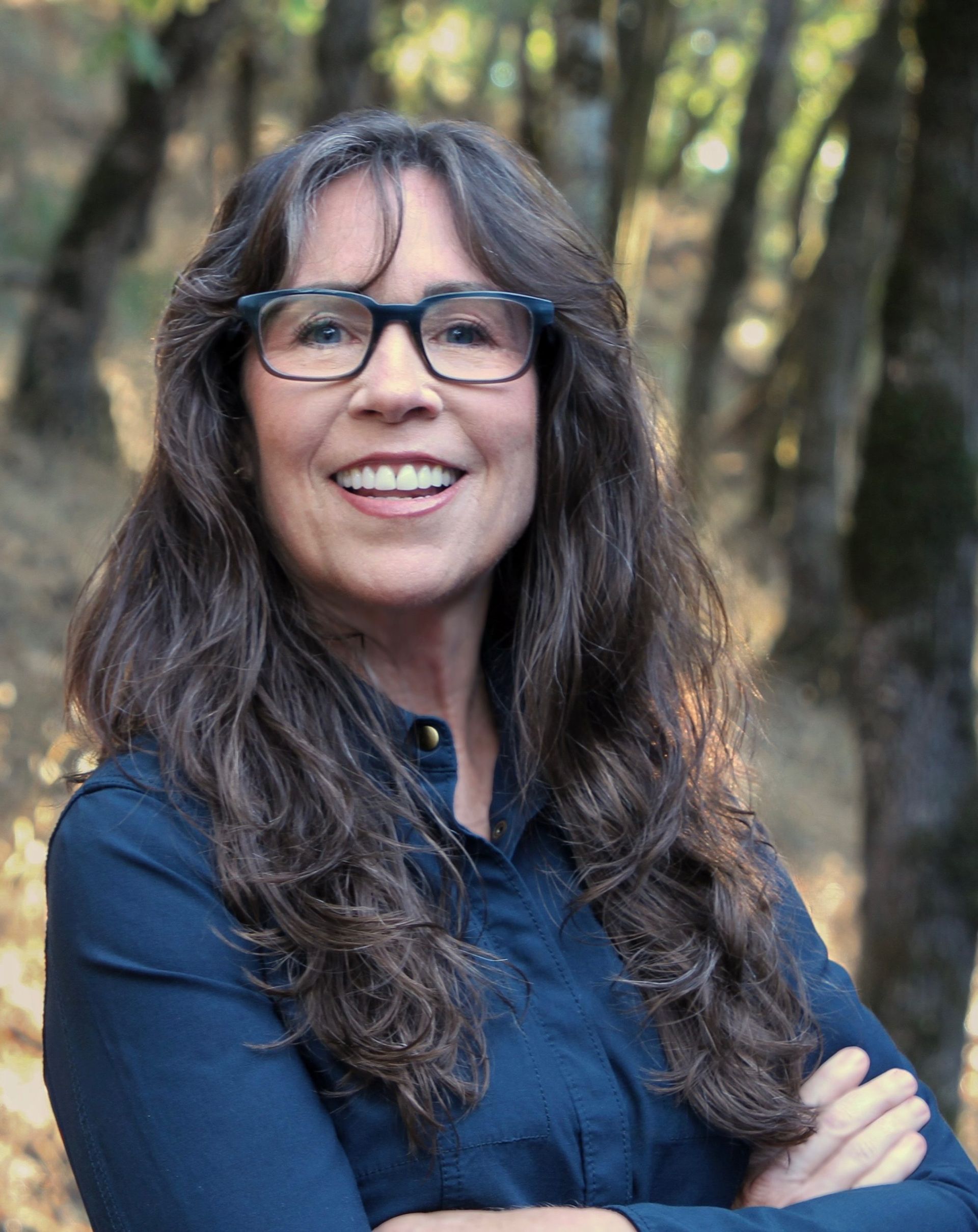 Woman smiling, wearing glasses, outdoors in front of blurred trees.