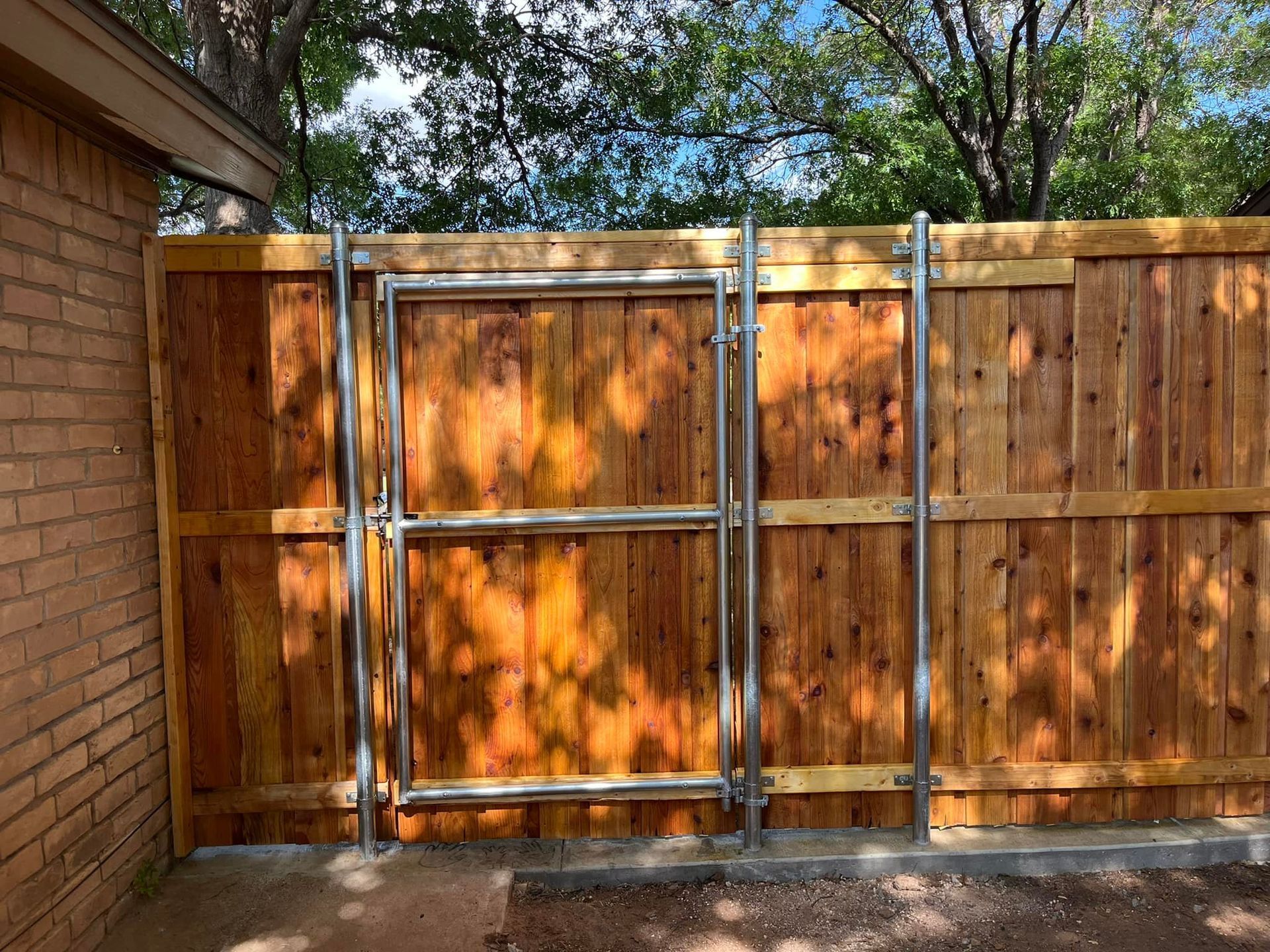 Wooden fence with metal gate, brick building on the left, blue sky visible through trees.