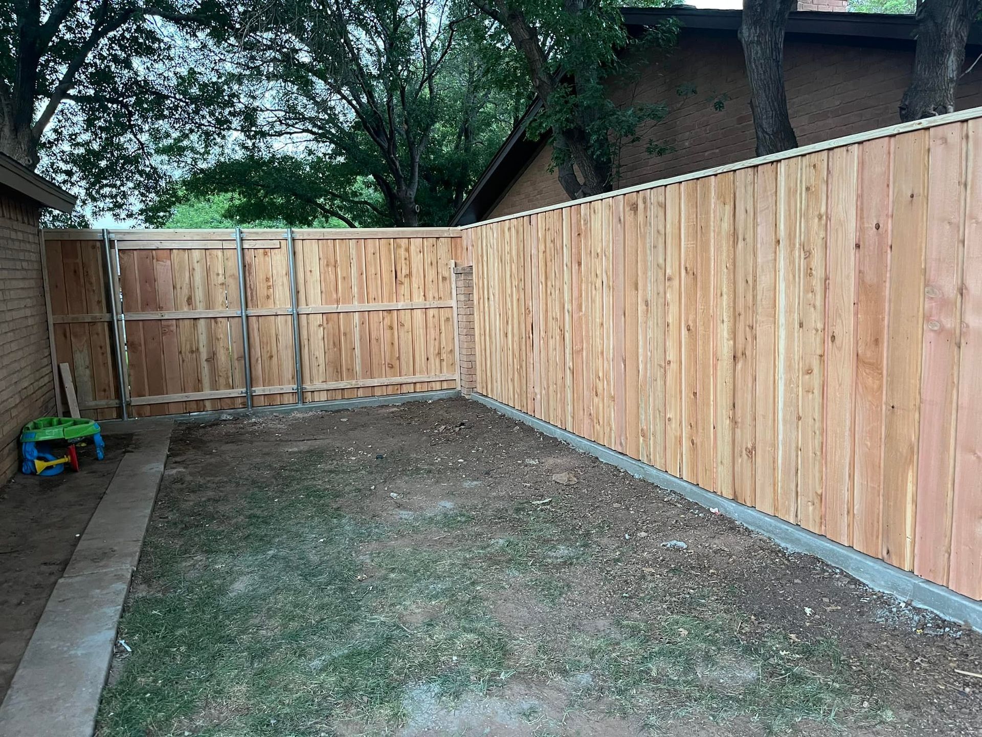 Wooden fence encloses a small backyard, with a brick wall, grass, and a few toys.