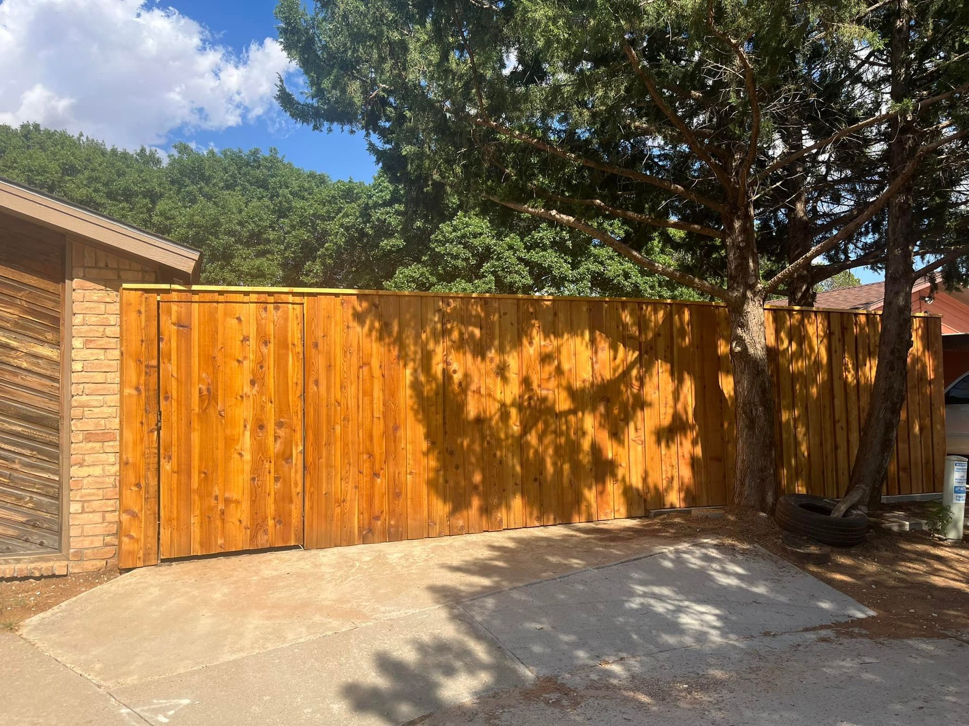 Wooden fence in front of a driveway.  A tree casts a shadow. Brown brick wall on the left.