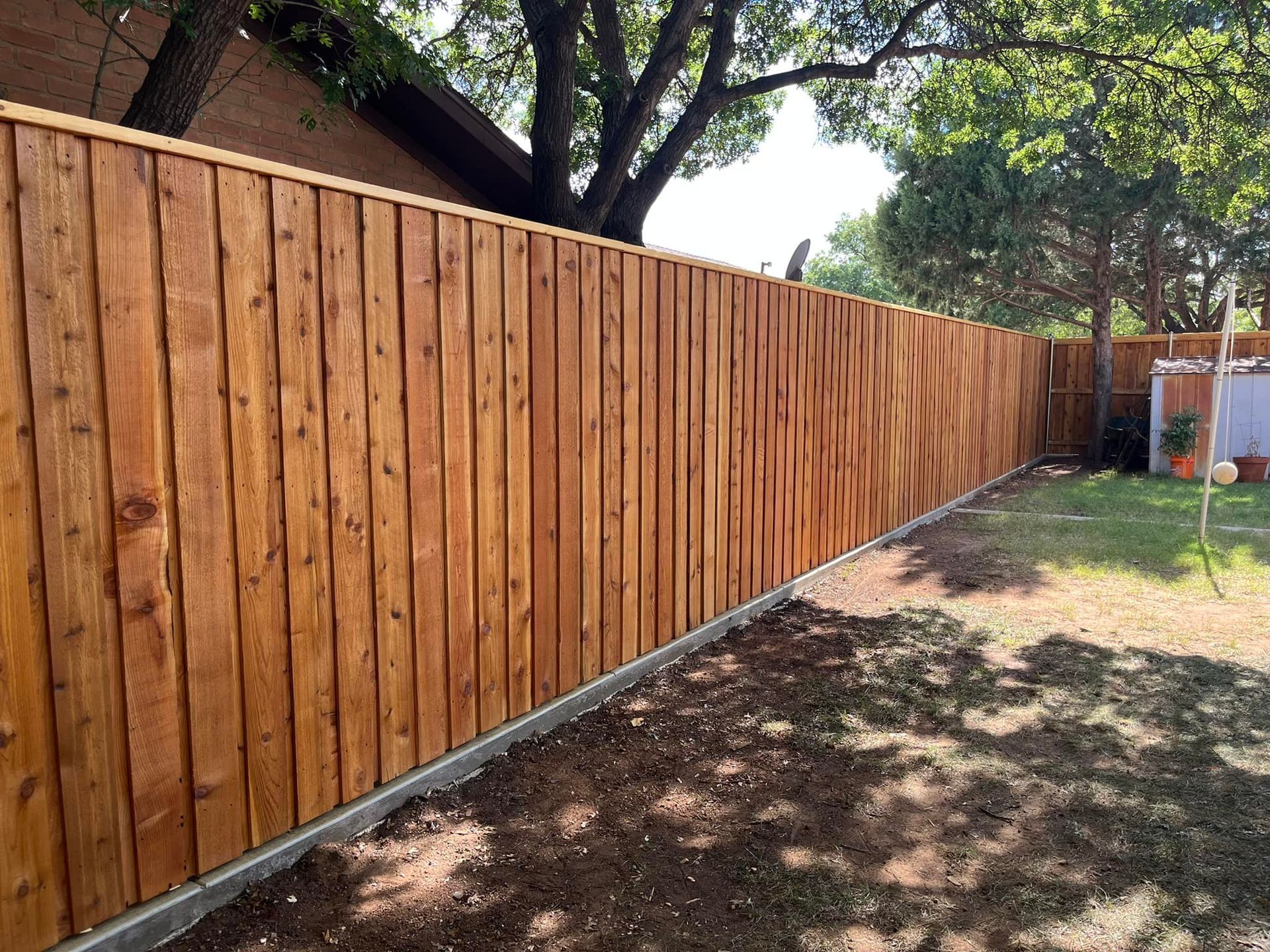 Wooden fence in backyard, brown wood planks, green grass, and trees.