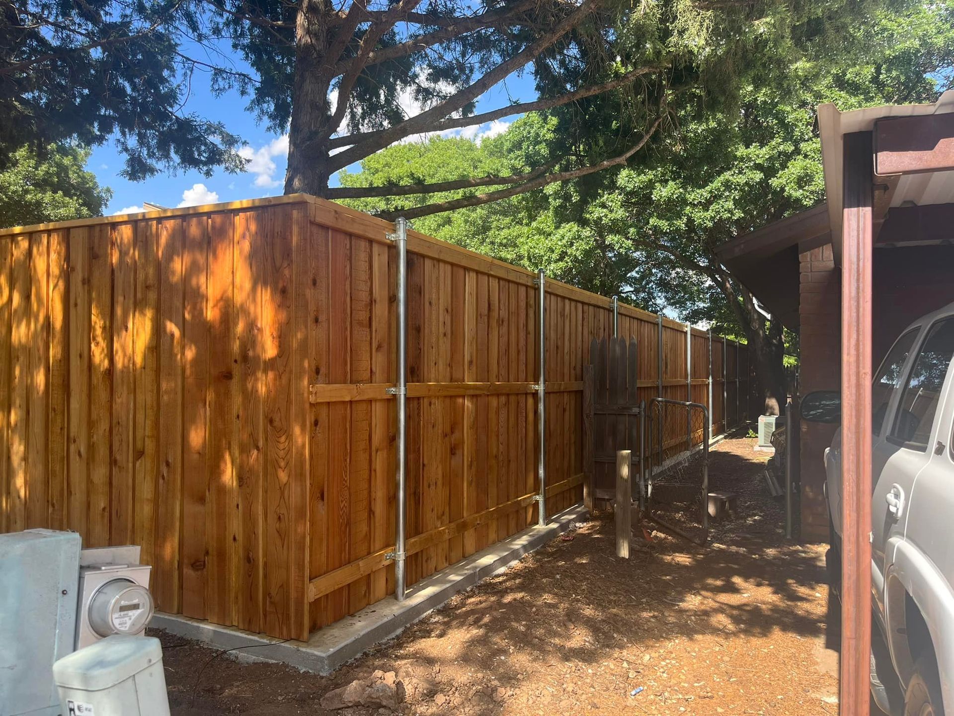Wooden fence in a yard, with metal posts. Brown stain. Sunny day, trees in the background.