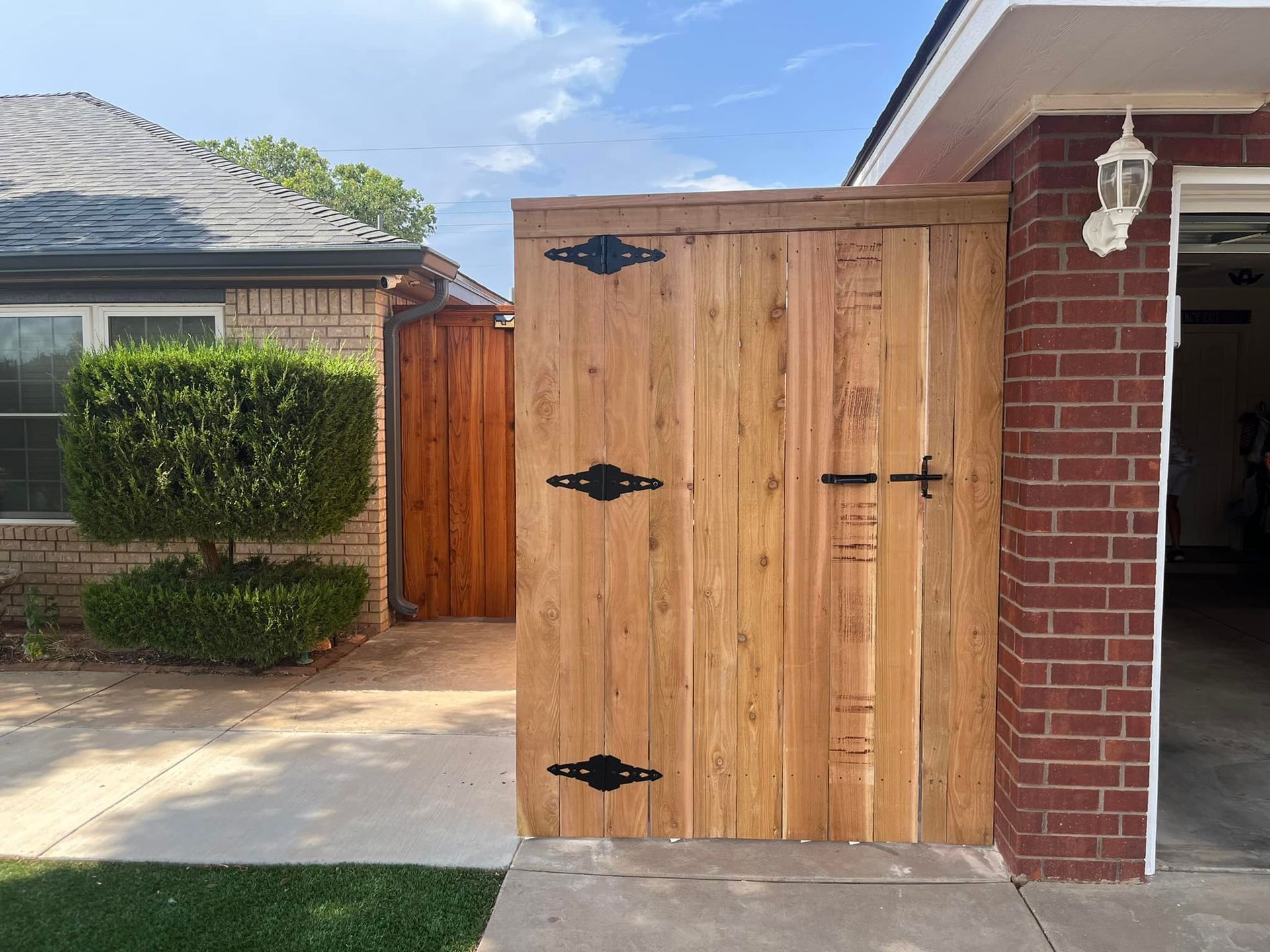 Wooden gate with black hardware, against a brick building and a driveway.