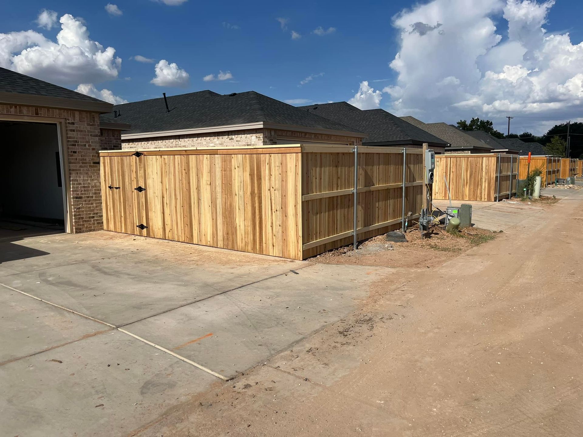 Wooden fences in a row of houses. Tan dirt road, concrete driveway, and blue sky.