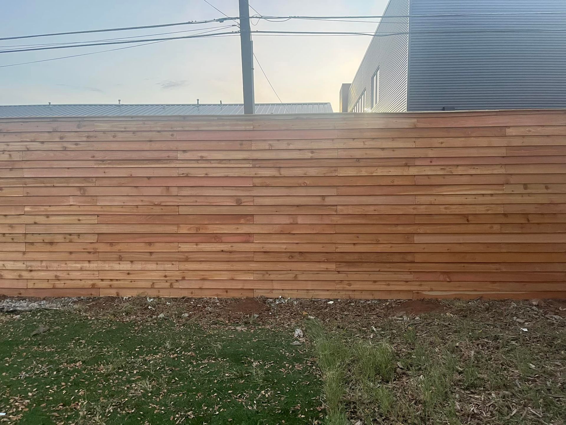 Wooden fence with a building featuring an optical illusion, in front of green grass and a partly cloudy sky.