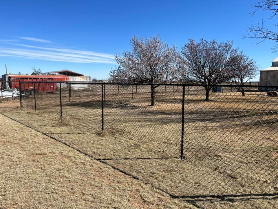 Black chain-link fence encloses a grassy area with leafless trees under a blue sky. Buildings are in the background.