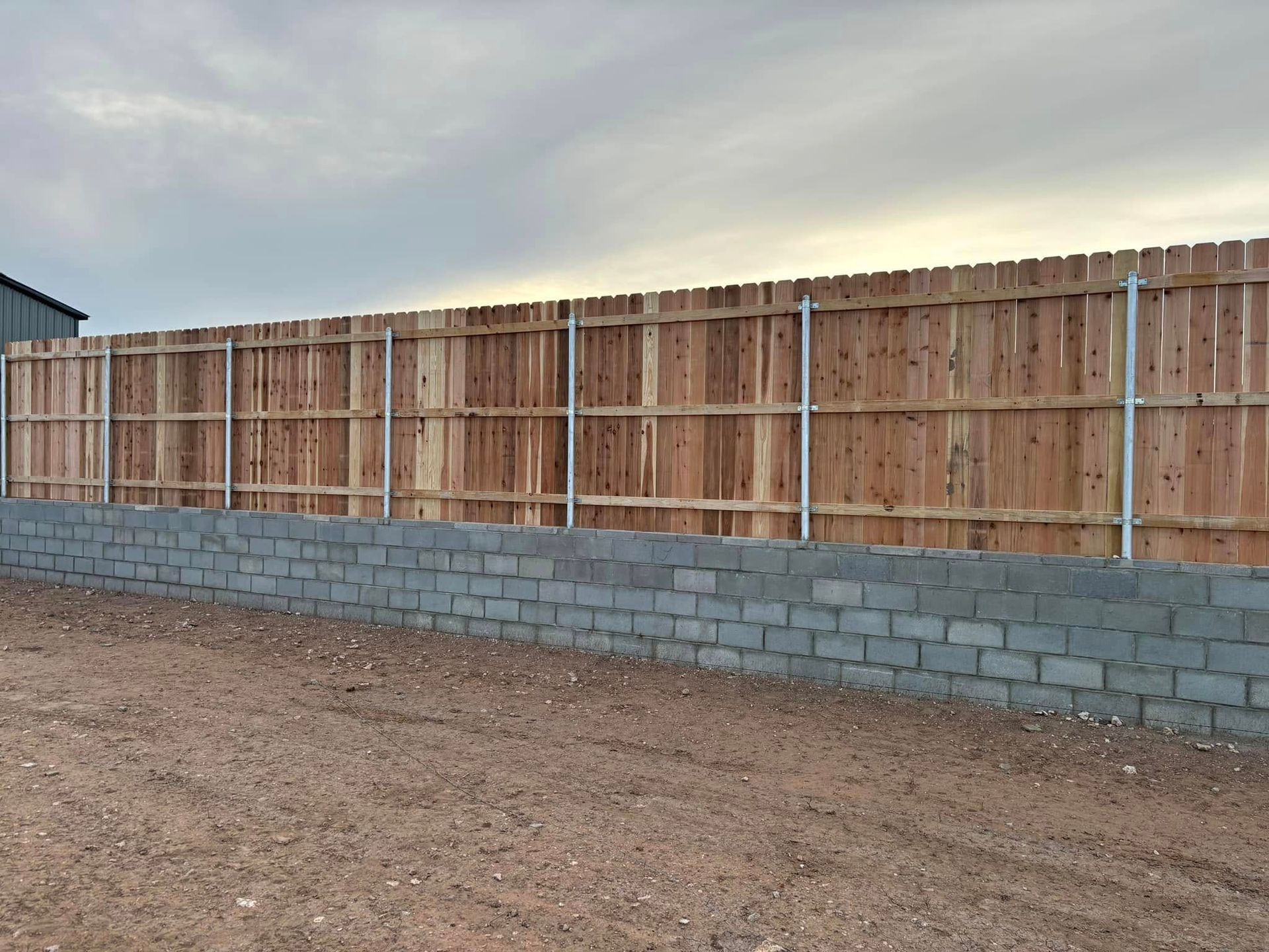 Wooden fence on a gray cinder block wall, set on a gravel ground under a cloudy sky.