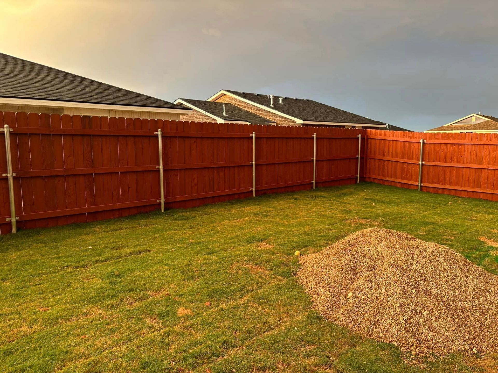Red wooden fence surrounds a grassy backyard with a pile of gravel. Overcast sky.