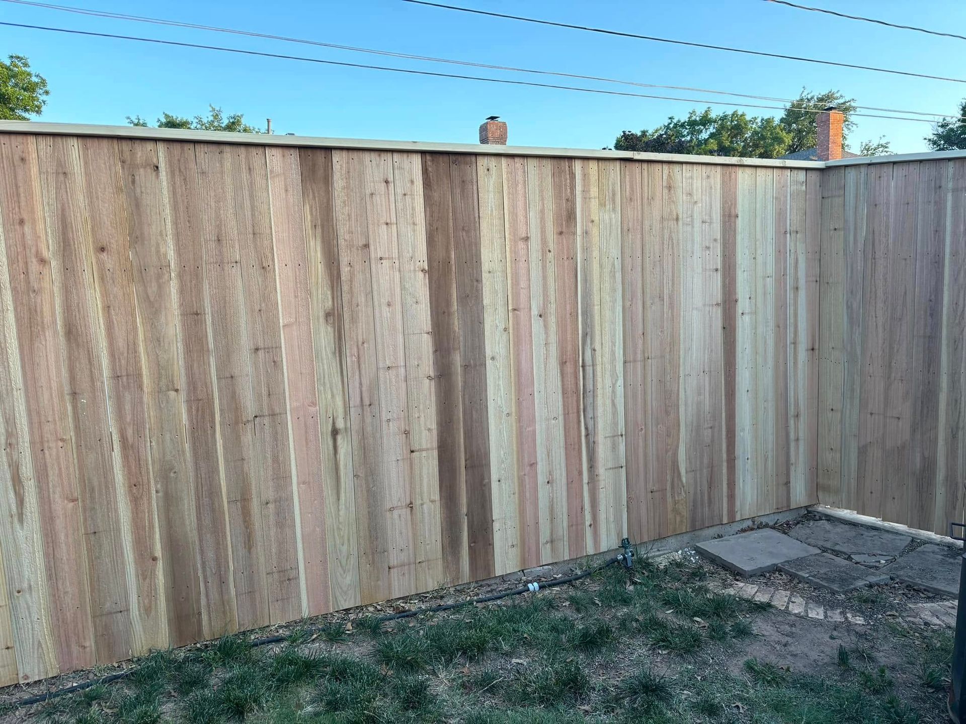 Wooden fence in a backyard with green grass, blue sky, and utility wires.