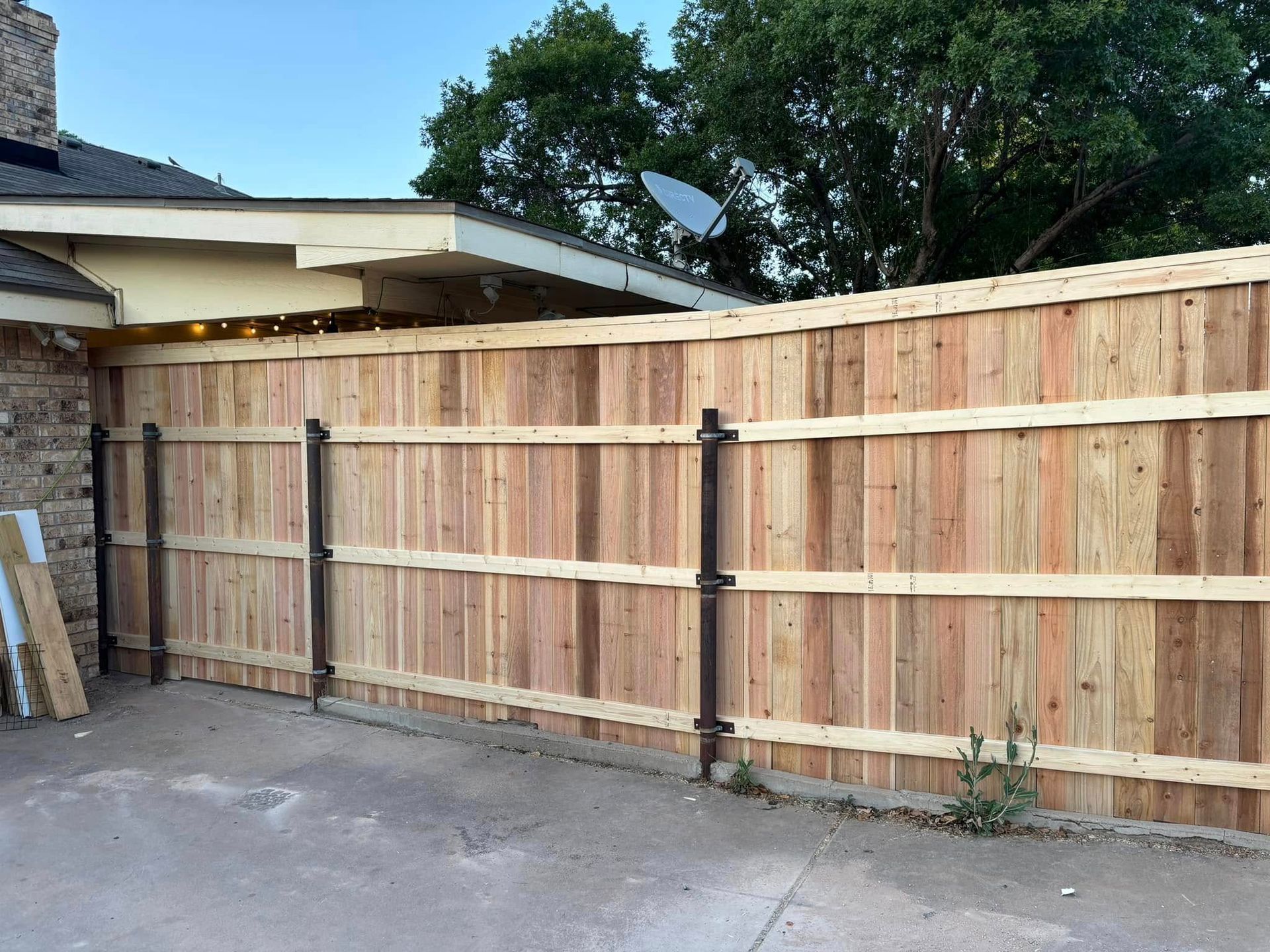 Wooden fence, built along a driveway, with a brown house, tree, and sky in the background.