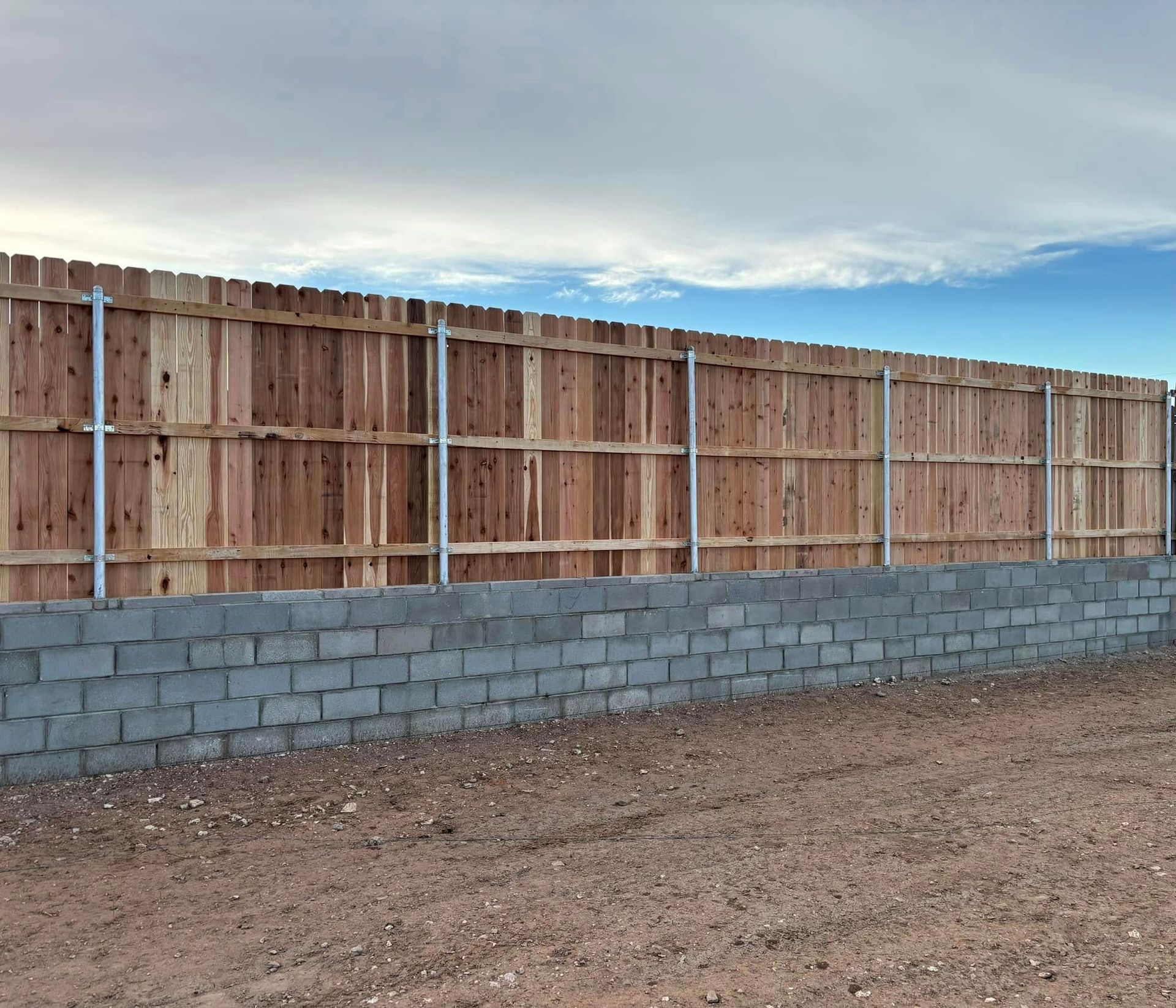 Wooden fence atop a cinder block wall, supported by metal posts. Sky and dirt in the background.