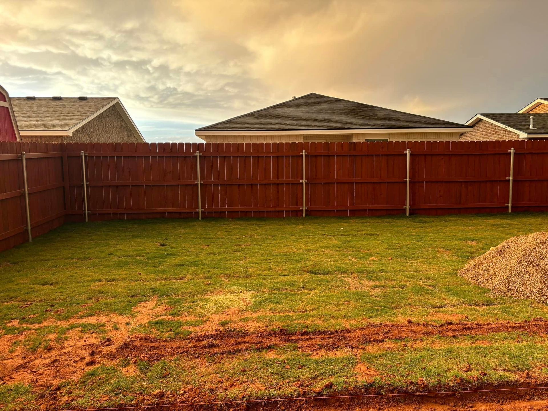Backyard with red wooden fence, green grass, and cloudy sky behind houses.