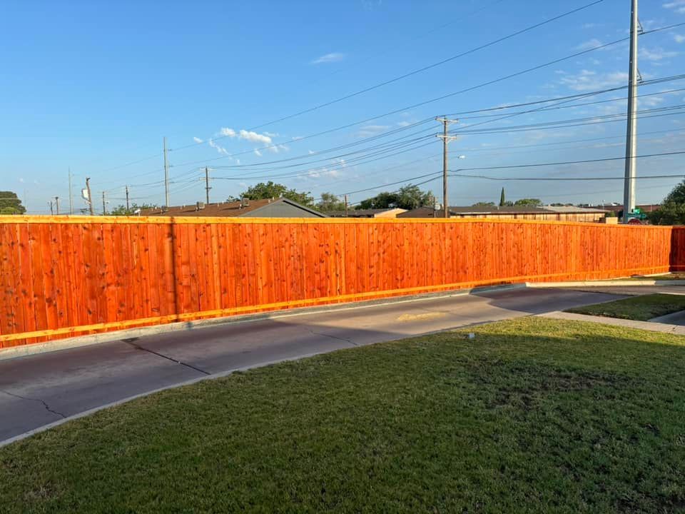 Red-stained wooden fence in front of a grassy lawn and paved road on a sunny day.