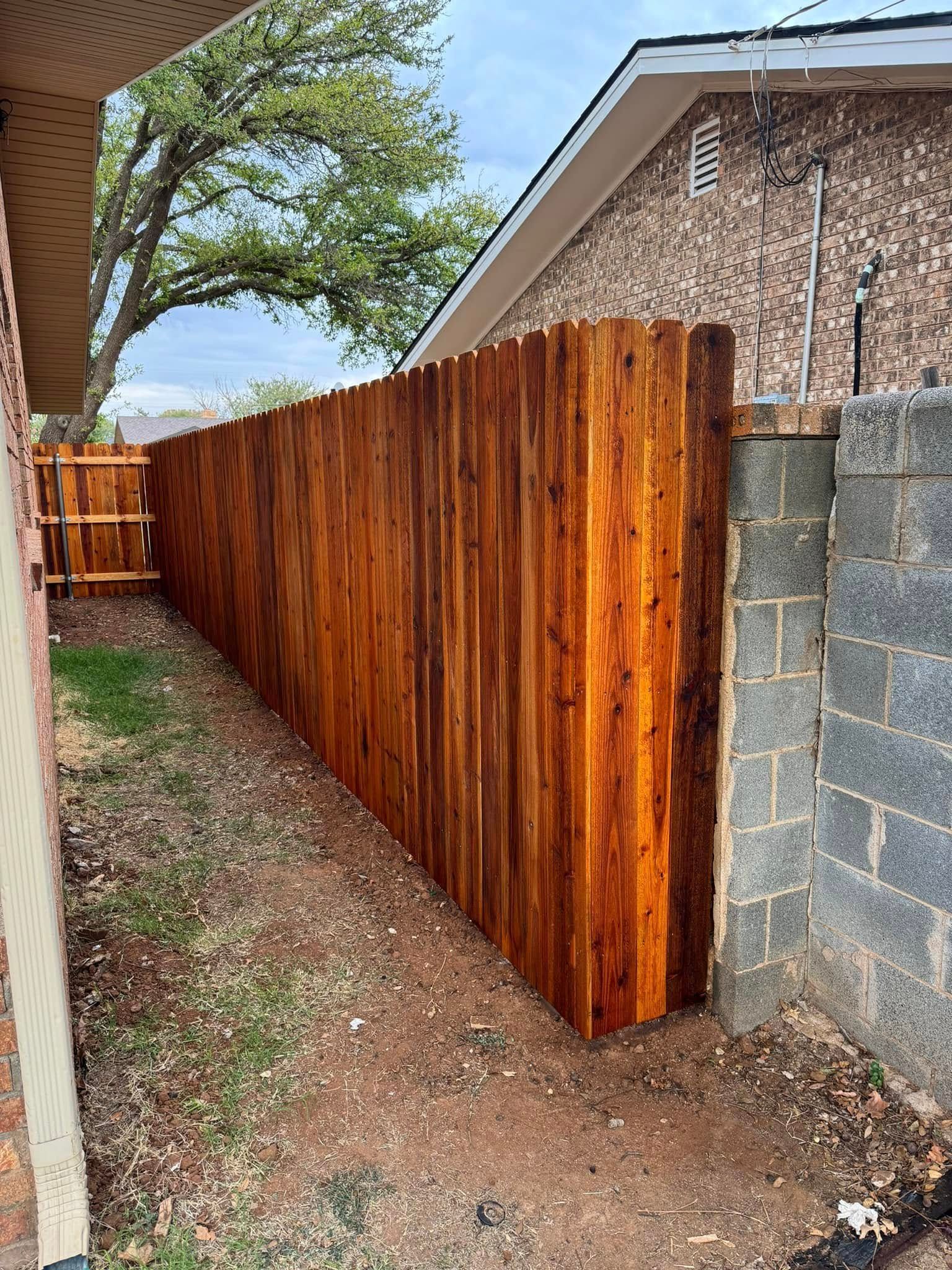 Wooden fence stained dark brown next to a brick wall and house; grass and bare earth in foreground.
