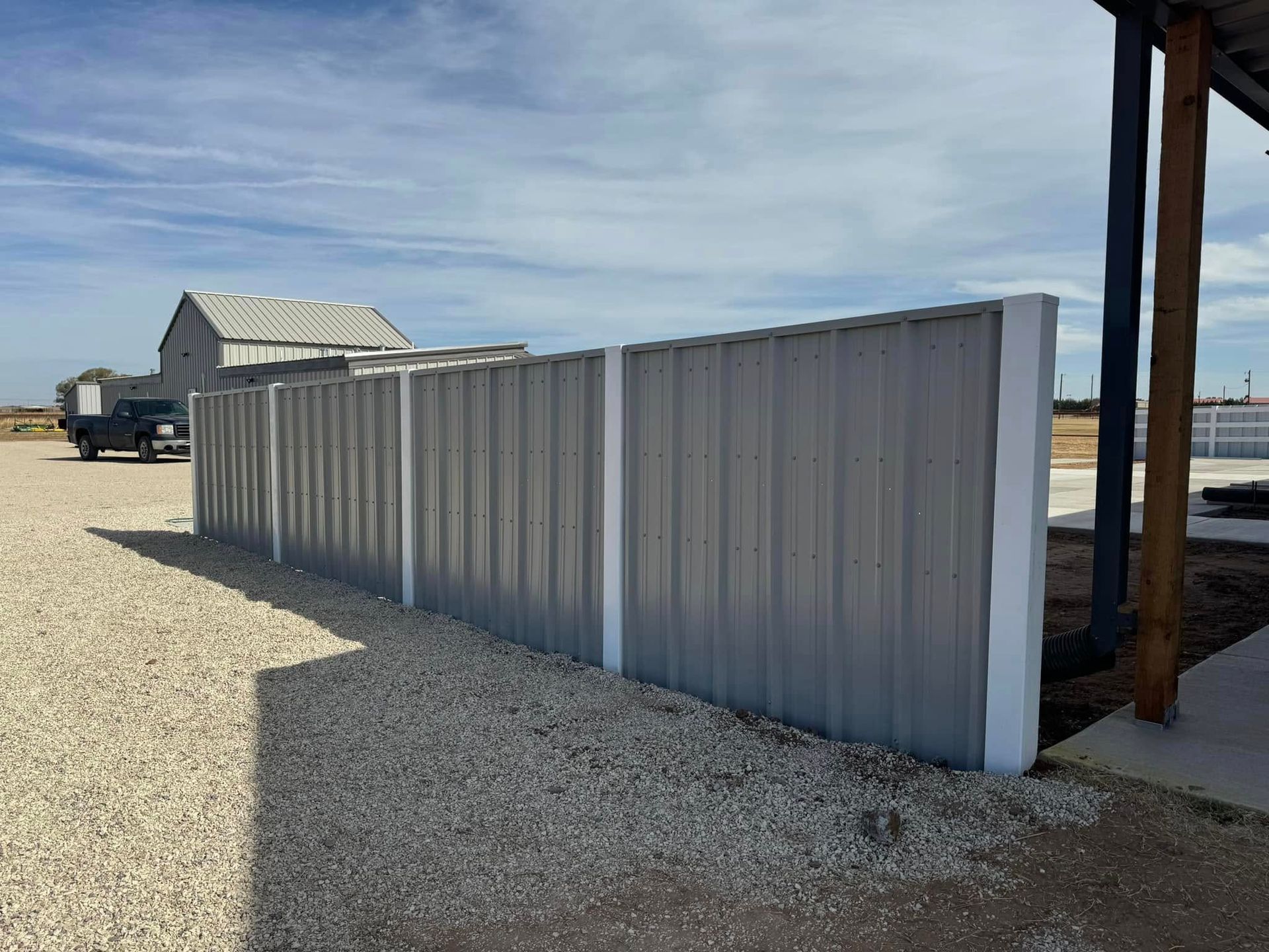 A metal fence with white posts on gravel in front of a barn under a blue sky.