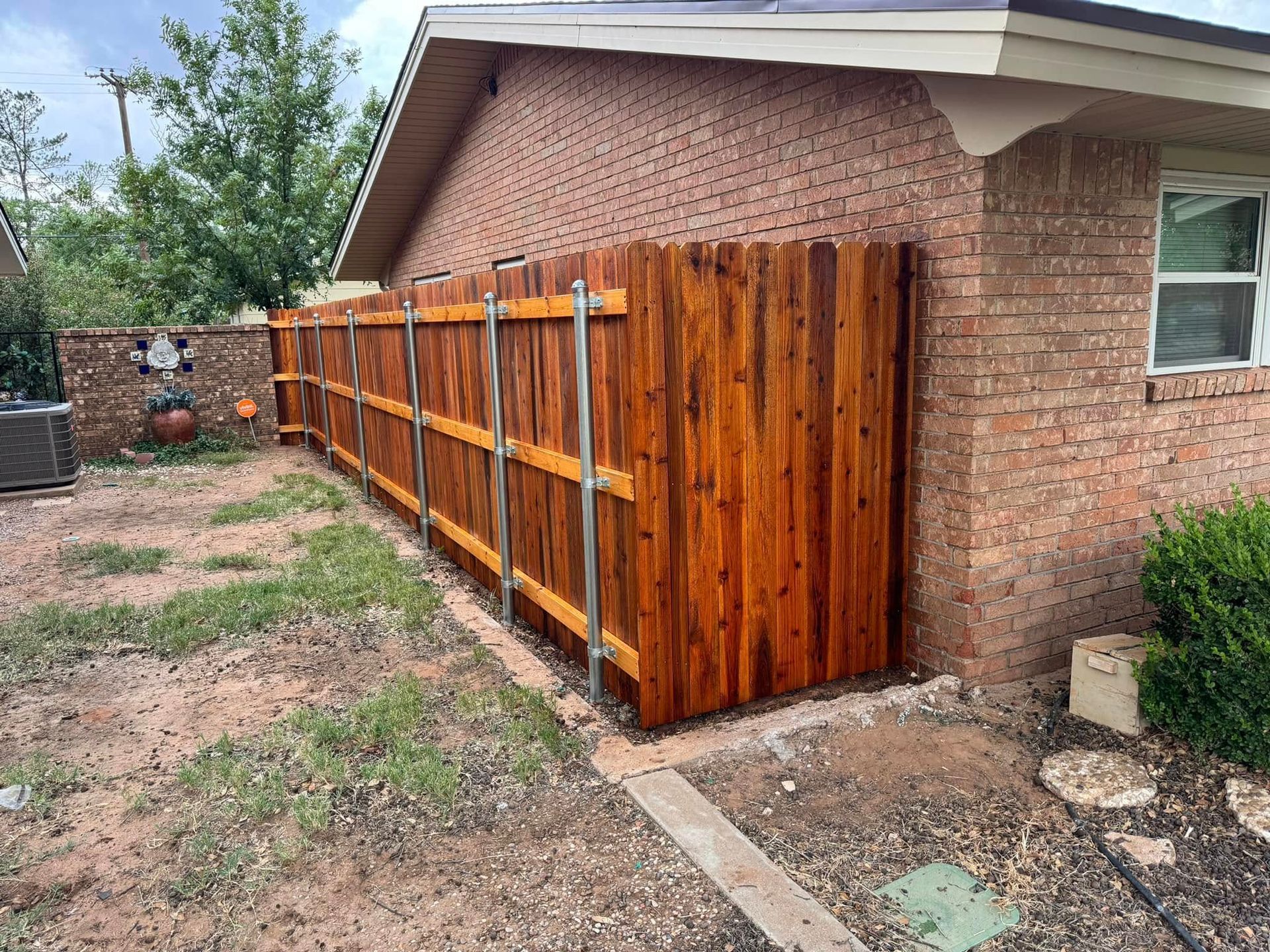 Wooden fence stained reddish-brown, attached to a brick house with silver supports. Outdoors, grass and a small bush visible.