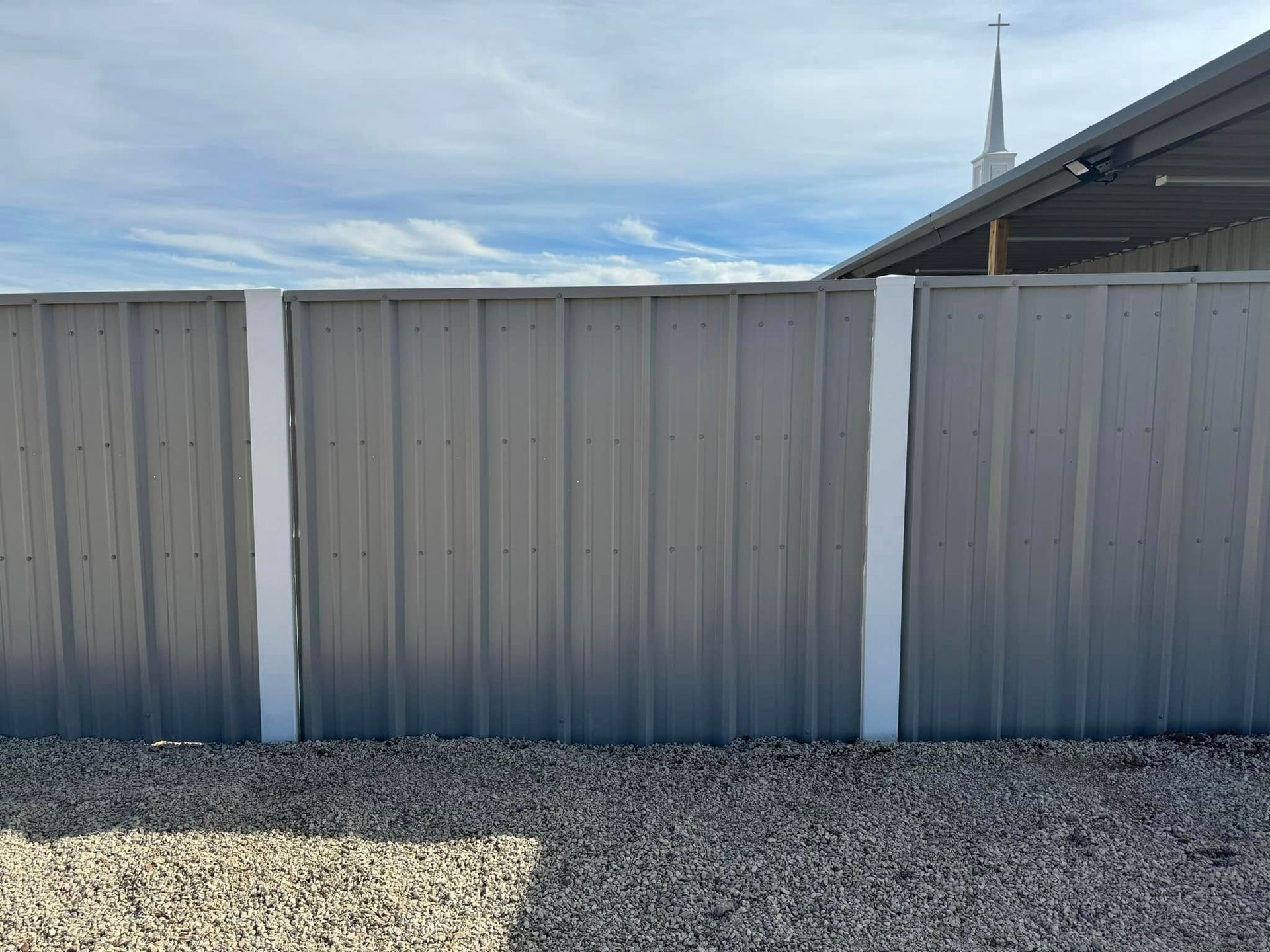 Gray metal fence with white posts against a blue sky, gravel ground, and a church spire.