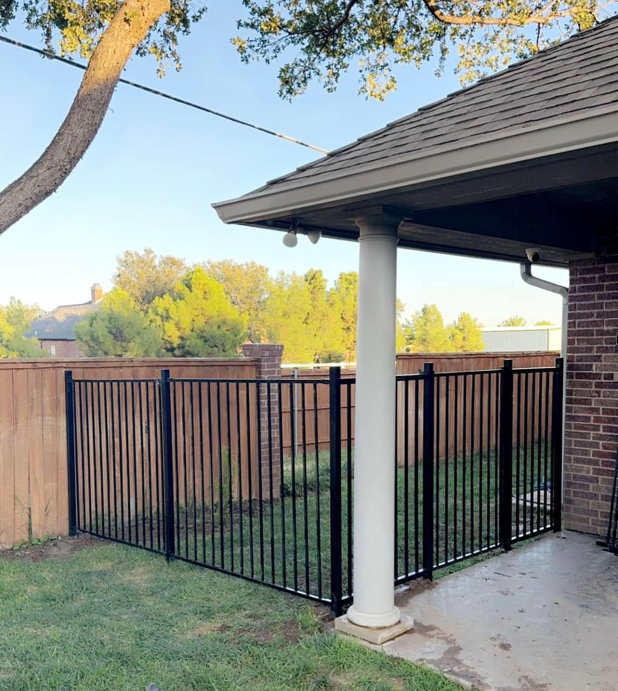 Black metal fence enclosing a backyard patio near a brick wall and wooden fence. Green grass, sunny day.